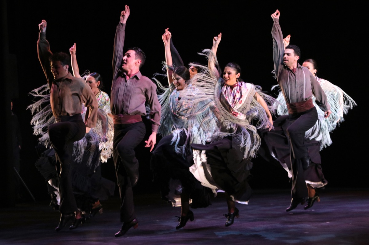 Grupo de hombres y mujeres bailando flamenco sobre un escenario, fondo negro.